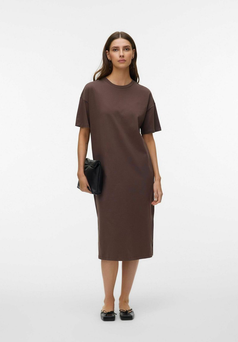 Femme debout portant une robe t-shirt midi marron ample, tenant une pochette noire et chaussée de ballerines noires.