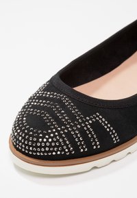 Sort flad sko med en blød, mat overflade, prydet med et sølvnagle-mønster på tåen og en let, holdbar sål.