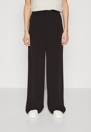 Person som har på seg svarte vide bukser og hvite joggesko, står foran en ensfarget lys bakgrunn.