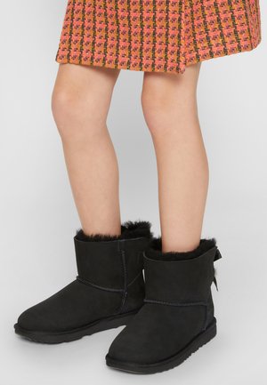 Bottines en peau de mouton noir avec doublure en fourrure, semelles plates et détail de couture sur le côté. Le design présente une texture douce et une silhouette basse.