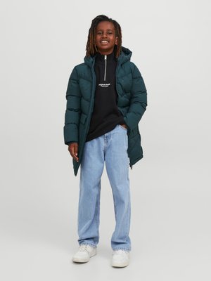 Lapsi, jolla on päällään tummanvihreä untuvatakki, musta vetoketjullinen huppari, vaaleansiniset farkut ja valkoiset tennarit, seisoo yhdellä kädellä taskussa.