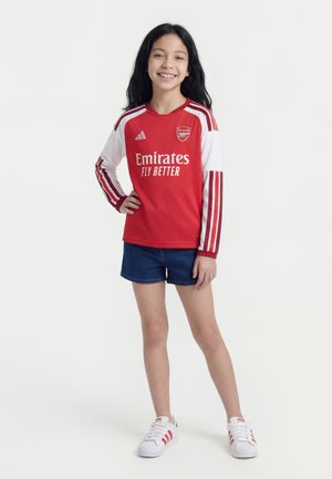 Niña sonriente de pie con la mano en la cadera, vestida con la camiseta de fútbol roja y blanca del Arsenal, pantalones cortos vaqueros y zapatillas blancas Adidas.