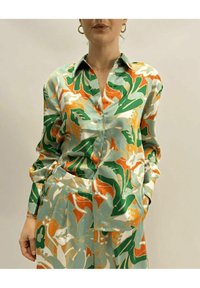 Camicia in satin verde, arancione e crema con motivo floreale; presenta un colletto, chiusura con bottoni e maniche lunghe con tasca.