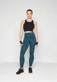 Sutiã desportivo preto, leggings de cintura alta em verde-azulado com um padrão subtil de camuflagem, luvas de fitness pretas e ténis pretos.