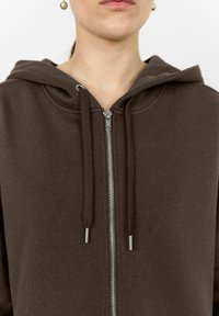 Felpa con zip marrone realizzata in tessuto morbido, dotata di cappuccio, zip argentata e cordini con punte metalliche. Design semplice e vestibilità casual.