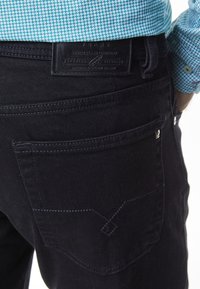 Primer plano de una persona que lleva puestos unos pantalones vaqueros negros con un bolsillo trasero visible y una camisa azul con estampado metida por dentro.