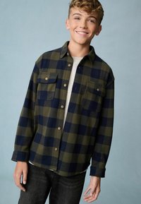 Flanellen overhemd in groen en marineblauw geruit, met twee borstzakken, knoopsluiting aan de voorkant en een relaxte pasvorm. Gekocht boven een lichtgrijs shirt.