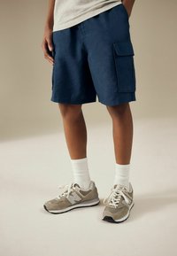 Pantaloni cargo blu navy in tessuto leggero con due tasche laterali, abbinati a scarpe da ginnastica grigie e calze bianche alla caviglia. Texture morbida e vestibilità rilassata.