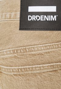 Denim beige con costuras en contraste, que presenta una etiqueta negra con el logotipo "DRDENIM". Superficie simple y texturizada con un detalle mínimo.