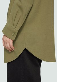 Olijfgroene lange mouwen shirt met een knoopmanchet en zijsplitten, gemaakt van een gestructureerde stof. Heeft een label nabij de zoom.