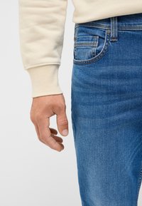 Beige sweatshirt med åtsittande mudd, bredvid blå denimjeans med en framficka med synlig söm och texturerad tyg.
