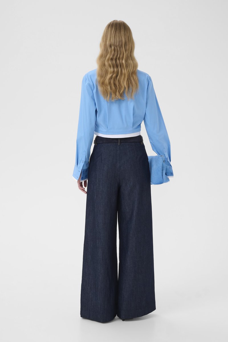Femme aux cheveux longs et ondulés portant une chemise bleu clair boutonnée et un pantalon large bleu foncé, tenant une pochette duveteuse bleue assortie.