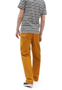 Person som bär en svartvit randig skjorta, senapsgula byxor och svarta slip-on skor, går bort från en vit bakgrund.