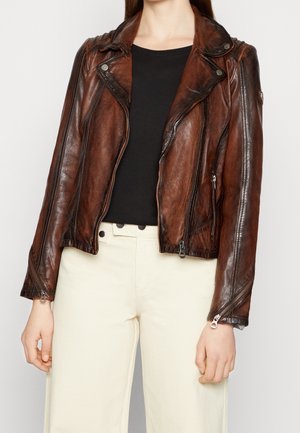 Kvinde iført en slidt brun læder bikerjakke over en sort top og lyse beige højttaljede bukser, stående mod en ensfarvet baggrund.