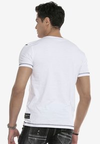 Weißes T-Shirt mit kurzen Ärmeln und schwarzen Nähten sowie zwei schwarzen Schulterklappen. Aus weichem, leichtem Stoff und figurbetonter Passform.