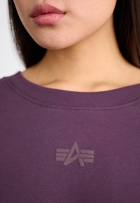 Fialová mikina s širokým výstřihem a texturovaným logem v světlejší fialové barvě, ukazující jemné švy na límci.