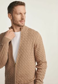 Beżowy kardigan z dzianiny z splotem warkoczowym, z suwakiem z przodu i wysokim kołnierzem. Prążkowane mankiety i dół dodają tekstury do projektu.