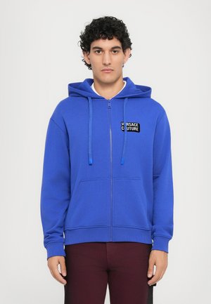 Blå lynlåshoodie lavet af blødt stof, med et logo patch på forsiden, snore og kænguru-lommer. Strukturerede ribkanter.