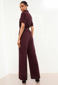 Mono burdeos con mangas cortas, cintura ceñida con cinturón, piernas anchas, tejido texturizado y bolsillos en el pecho con botones. Se lleva con tacones negros.