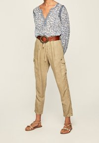 Blusa floreale azzurra chiara con scollatura a V, pantaloni cargo in lino beige, cintura marrone e sandali piatti con cinturini decorativi. Look texture e informale.