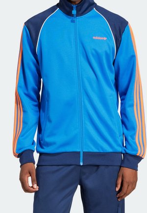 Veste de survêtement Adidas bleu et marine zippée avec des bandes orange sur les manches portée par une personne portant un pantalon foncé.
