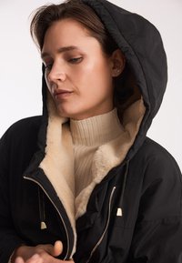 Czarna kurtka z kapturem, z miękkim beżowym wykończeniem, z zapięciem na zamek i złotymi akcentami metalowymi. Pod spodem sweter z ryżu.