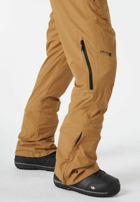 Tan sneeuwbroek met een rechte snit, zijzakken met rits en zwarte hardware. Gecombineerd met zwarte sneeuwlaarzen met een gestructureerde zool.