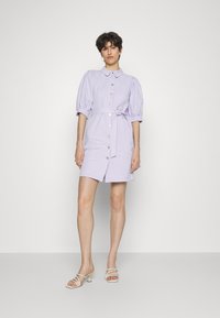 Vestido curto de cor lilás, com mangas curtas, gola, fecho à frente com botões, cintura ajustada e um cinto atado. Tecido suave com um design discretamente estruturado.