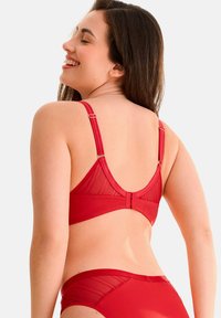 Soutien-gorge rouge avec un design en tissu maillé et solide, bretelles réglables avec des détails en métal doré, et une fermeture à crochets.
