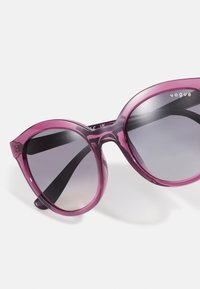 Ochelari de soare rotunzi, supradimensionați, cu un cadru translucid de culoare roz închis, lentile gri cu gradient și brațe negre. Logo-ul "Vogue" pe partea superioară a cadrului.