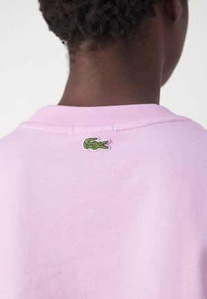 Vaaleanpunainen puuvillainen collegepaita pyöreällä kaula-aukkolla ja resorikauluksella, jossa pieni vihreä krokotiililogo brodeerattuna takana.