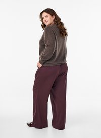 Pull en tricot marron à épaules tombantes, associé à un pantalon ample de couleur bordeaux foncé. Le mannequin pose avec une main dans la poche.