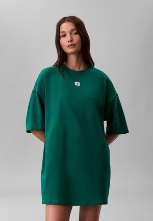 Kobieta z długimi brązowymi włosami, ubrana w luźną, ciemnozieloną, dużą sukienkę typu T-shirt z małą białą naszywką z logo na piersi, stojąca na tle jednolitego szarego tła.