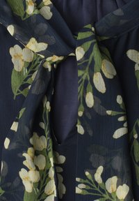 Tissu transparent foncé à imprimé floral blanc et jaune noué en un nœud sur un fond uni bleu foncé.