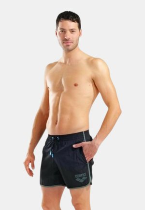 Uomo in piedi senza maglietta, indossa pantaloncini da bagno neri con coulisse e logo del marchio sulla gamba sinistra, mano in tasca.