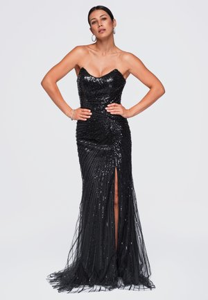 Femme debout, mains sur les hanches, portant une robe bustier noire à sequins avec une fente devant et des boucles d’oreilles créoles sur un fond clair uni.
