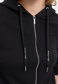 Čierna mikina na zips s mäkkou tkaninou, kapucňou na šnúrku, strieborným zipsom a vyšitým logom na hrudi.
