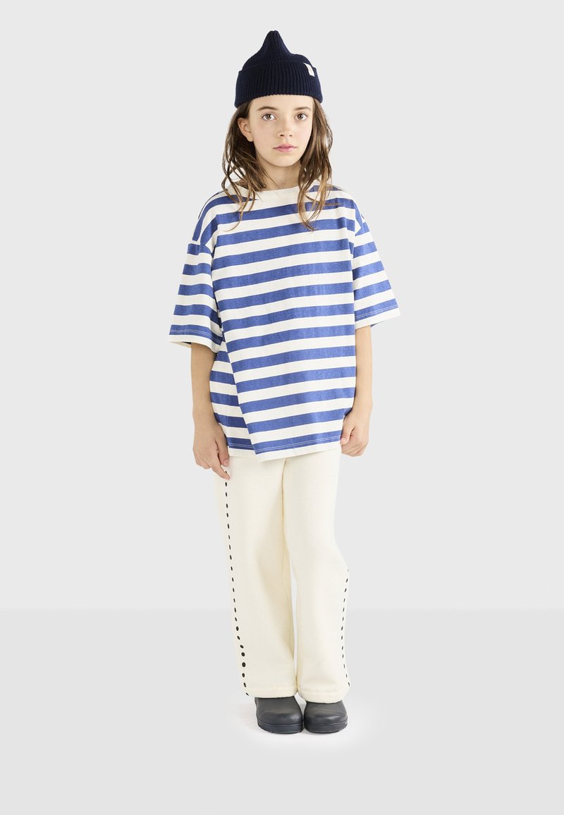 Stribet blå og hvid oversized t-shirt, cremefarvede bukser med sort stikning i siderne, og en marineblå hue. Sorte sko fuldender outfittet.