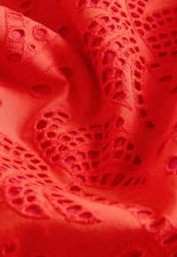 Tissu rouge avec broderie œillets complexe et bords festonnés créant un motif texturé et décoratif.