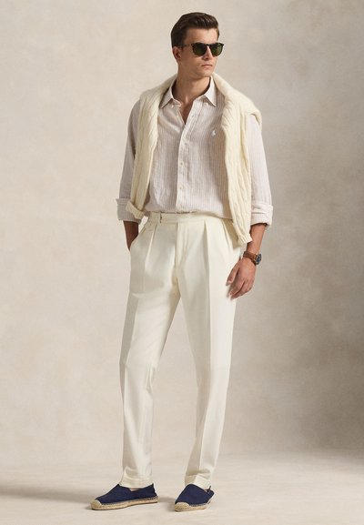Homme portant des lunettes de soleil, une chemise claire à rayures, un pantalon crème, des espadrilles bleu marine et un pull en tricot crème drapé sur les épaules.