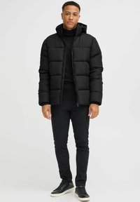 Black puffer jacket with a hood, featuring quilted sections, zip closure, and a soft texture. Paired with black skinny pants and sneakers.