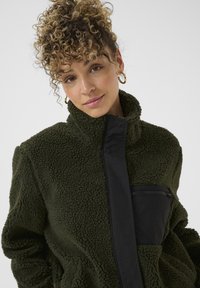 Groene fleecejack met hoge kraag, zwarte ritssluiting zak en gestructureerd materiaal. Heeft een relaxte pasvorm en een zachte oppervlakte.