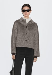 Brown, cropped wool jacket with a collar, button closure, and long sleeves, paired with a black turtleneck and pants, held with a bag.