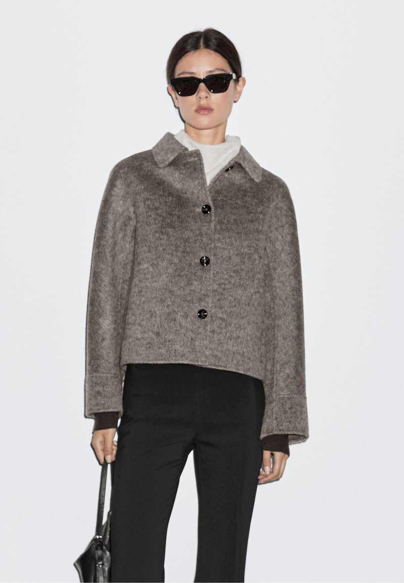 Brown, cropped wool jacket with a collar, button closure, and long sleeves, paired with a black turtleneck and pants, held with a bag.