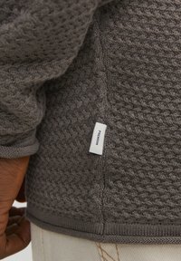 Suéter gris de punto con un patrón texturizado, que presenta una etiqueta blanca con "PREMIUM". Puños y dobladillo elásticos, destacando un corte relajado.