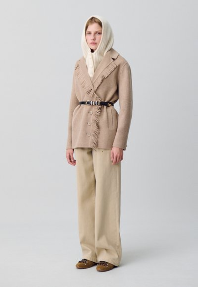 Femme portant un manteau à franges beige ceinturé à la taille, un pull à capuche crème en maille, un pantalon large beige et des chaussures marron, se tenant sur un fond gris uni.
