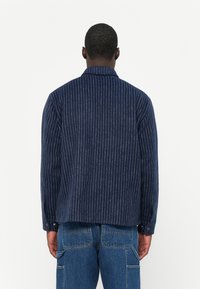 Dunkelblaue Jacke mit weißen vertikalen Streifen, ausgestattet mit einem Kragen und langen Ärmeln. Weicher Stoff mit strukturierter Oberfläche und Knopfakzenten.