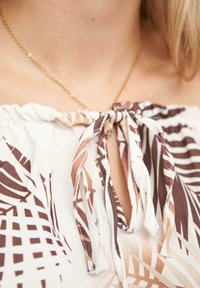 Blusa holgada con diseño fuera del hombro, que presenta un patrón geométrico en marrón y beige, escote atado y textura de tela suave.