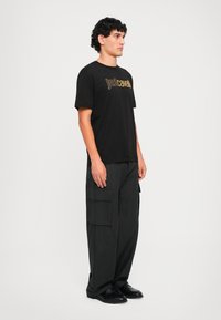 Melna īsām piedurknēm T-kreklā ar zelta "Just Cavalli" logotipu, apvienota ar brīviem melniem kravas biksēm ar vairākām kabatām.