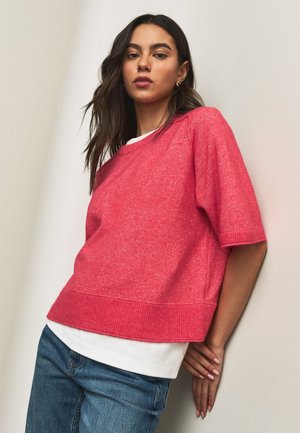 Jeune femme en pull rose à manches courtes sur chemise blanche et jean bleu, appuyée contre un mur clair, mains jointes dans le dos.